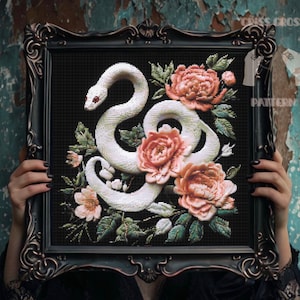 May include: A framed cross-stitch pattern featuring a white snake with red eyes, intertwined with pink and peach-colored flowers and green leaves, set against a black background. The frame is ornate and black. The words "CROSS CROSS PATTERN" are visible.