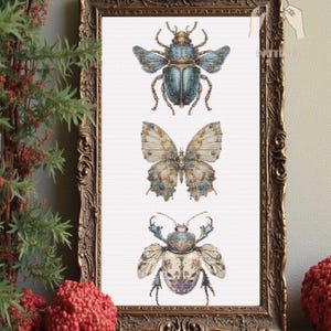 May include: A framed artwork featuring cross-stitch designs of three insects: a beetle, a butterfly, and another beetle. The insects are in shades of blue, beige, and brown, set against a white background. The frame is ornate with a gold finish.