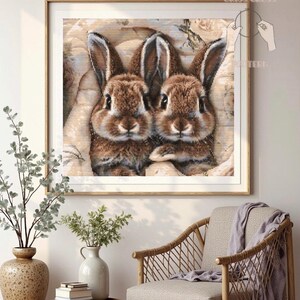 May include: Framed cross-stitch art featuring two brown rabbits with long ears and detailed fur. The artwork is set against a soft, neutral background and framed in a light wood. The text "Criss Cross Pattern" is visible.