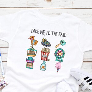 May include: White t-shirt with the text "TAKE ME TO THE FAIR" and colorful illustrations of carnival rides and treats. The shirt is paired with white sneakers and colorful markers on a wooden surface.