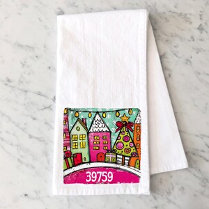 May include: A white kitchen towel featuring a colorful illustration of a winter village scene with houses, a decorated Christmas tree, and gift boxes under a string of lights. The number 39759 is printed in white on a pink curved banner at the bottom.