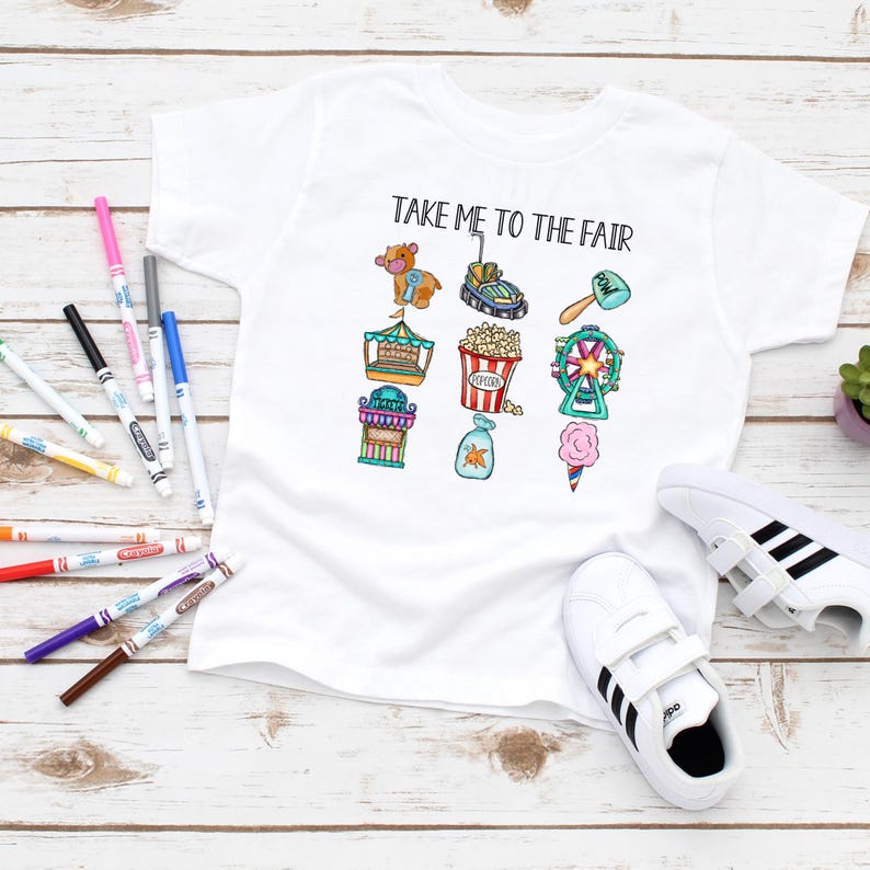 May include: White t-shirt with the text "TAKE ME TO THE FAIR" and colorful illustrations of carnival rides and treats. The shirt is paired with white sneakers and colorful markers on a wooden surface.