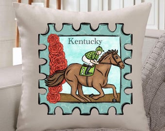 May include: A beige throw pillow featuring a horse racing design. The design includes a brown horse with a jockey, a row of red roses, and the word "Kentucky" within a blue stamp-like frame.