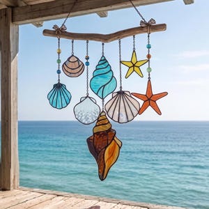 Puede incluir: Móvil de vidrieras con diseños de conchas marinas y estrellas de mar en tonos azules, naranjas, amarillos y blancos. El móvil está suspendido de una rama de madera con detalles de cuentas, creando un tema costero. El fondo muestra el mar y el cielo.