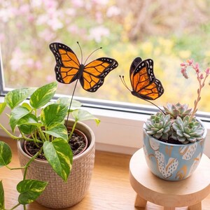 May include: Two monarch butterfly garden stakes with orange and black stained glass wings. One butterfly is positioned in a textured beige pot with a green pothos plant, and the other is in a light blue patterned pot with a succulent plant.