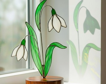 May include: A stained glass snowdrop sculpture with white flower petals and green leaves. The sculpture is mounted on a round, dark wooden base. The sunlight casts a shadow of the snowdrop on the wall.