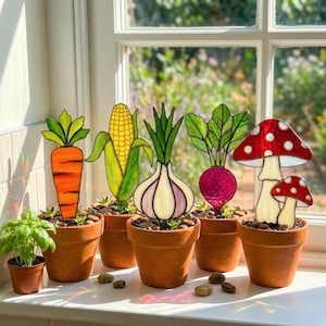 Puede incluir: Adornos de jardín de vidrieras que representan una zanahoria, maíz, cebolla, remolacha y setas. Cada pieza está colocada en una maceta de terracota con pequeñas piedras, creando una exhibición caprichosa. Los colores vibrantes y diseños detallados son encantadores.