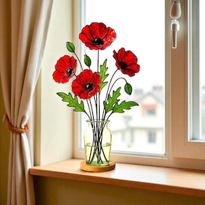 Pode incluir: Uma peça de arte em vitral com quatro flores de papoula vermelhas vibrantes com centros pretos e folhas verdes, apoiadas por hastes de metal preto finas. As flores estão dispostas em um vaso de vidro transparente sobre uma base de madeira.