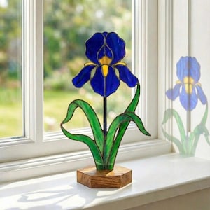 May include: A stained glass iris flower sculpture. The flower has deep blue petals with yellow accents, green leaves, and a wooden base. The sculpture is displayed on a windowsill, with a blurred background of greenery.