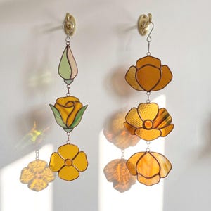 May include: Two vertical stained glass wall hangings suspended from brass hooks. The left piece features a pale green bud, a yellow rose with green leaves, and a yellow flower. The right piece displays three amber-colored floral shapes.