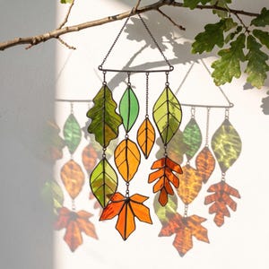 May include: A stained glass mobile featuring multiple leaf-shaped glass pieces in shades of green, yellow, and orange. The leaves are connected by thin metal chains to a horizontal metal bar, which is suspended by a chain from a tree branch.