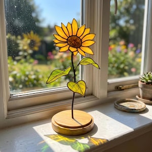 以下が含まれることがあります： 鮮やかな黄色の花びらと濃い茶色の中心部を持つステンドグラスのヒマワリの彫刻。2枚の緑の葉が付いた細い金属製の茎で支えられ、窓辺の明るい色の円形木製ベースに取り付けられています。