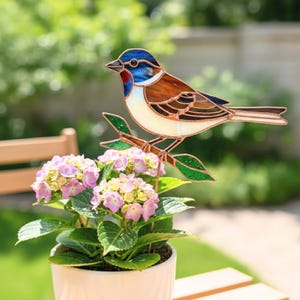 Può includere: Decorazione a forma di uccello in vetro colorato con dettagli blu, marroni, bianchi e rossi, posato su un ramo di rame con foglie verdi. L'uccello è posto in un vaso bianco con fiori rosa e gialli.