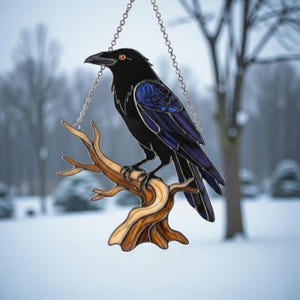 May include: A stained glass raven with black feathers and blue wing accents perches on a brown branch. The raven has orange eyes and hangs from a silver chain. The background is a snowy, blurred winter scene.