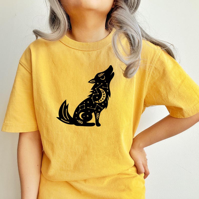 May include: Yellow t-shirt featuring a black graphic of a howling wolf with celestial details. The wolf design includes a crescent moon and stars. The shirt has short sleeves and a crew neck.