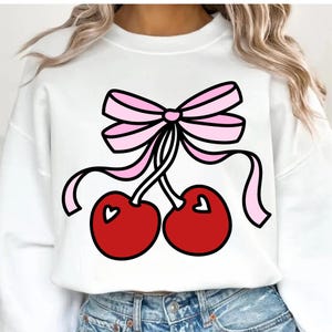 May include: White sweatshirt featuring a graphic of two red cherries with heart-shaped details, tied with a pink bow. The design is outlined in black. The sweatshirt is a casual, everyday garment.