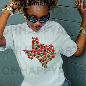 May include: A light blue t-shirt featuring a graphic of the state of Texas filled with red poppy flowers. The shirt is a crew neck style and is made of a soft material. The text "OH HAPPY ARTS" is visible in the background.