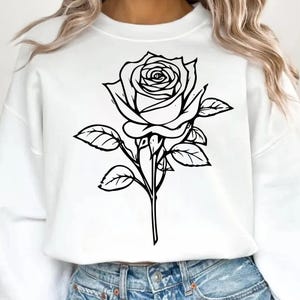 May include: White sweatshirt featuring a large black and white rose graphic. The rose is detailed with petals and leaves, creating a botanical design. The sweatshirt is a casual style, suitable for everyday wear.