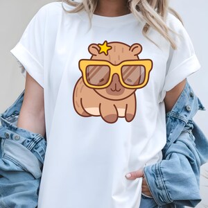K&ouml;nnte beinhalten: Wei&szlig;es T-Shirt mit einem Cartoon-Wasserschwein, das eine gelbe Sonnenbrille und einen Stern tr&auml;gt. Das Wasserschwein ist braun mit einem hellbraunen Bauch. Das T-Shirt hat hochgekrempelte &Auml;rmel und wird mit einer Jeansjacke kombiniert.
