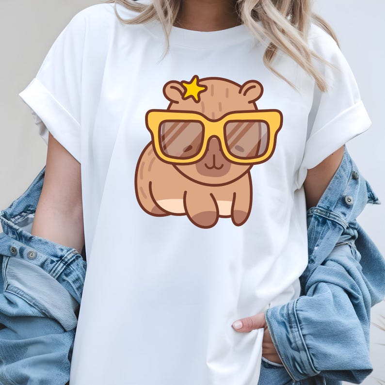 K&ouml;nnte beinhalten: Wei&szlig;es T-Shirt mit einem Cartoon-Wasserschwein, das eine gelbe Sonnenbrille und einen Stern tr&auml;gt. Das Wasserschwein ist braun mit einem hellbraunen Bauch. Das T-Shirt hat hochgekrempelte &Auml;rmel und wird mit einer Jeansjacke kombiniert.