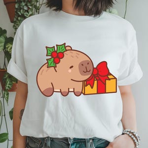 K&ouml;nnte beinhalten: Wei&szlig;es T-Shirt mit einem Cartoon-Capybara-Design. Das hellbraune Capybara tr&auml;gt Stechpalme und h&auml;lt ein gelbes Geschenk mit einer roten Schleife. Ein festliches und niedliches Design.