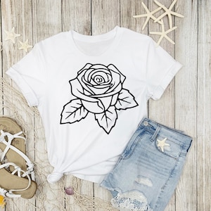 May include: White t-shirt featuring a large black outline of a rose with leaves. The shirt is paired with denim shorts and sandals, suggesting a casual, summery style. The design is simple and graphic.