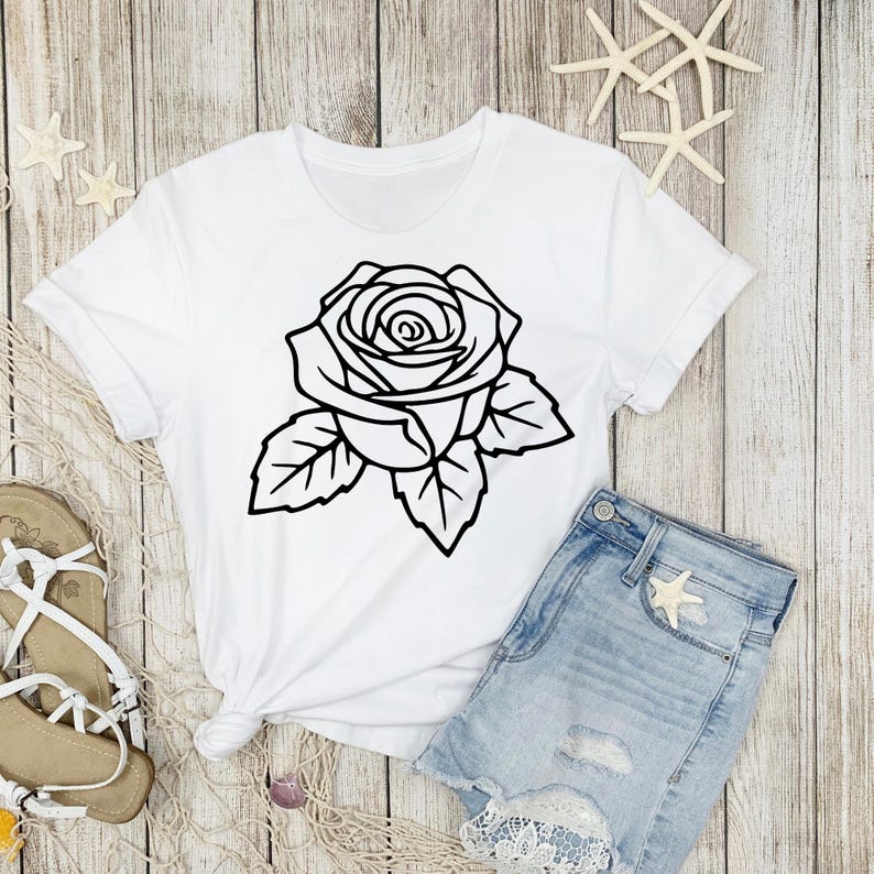 May include: White t-shirt featuring a large black outline of a rose with leaves. The shirt is paired with denim shorts and sandals, suggesting a casual, summery style. The design is simple and graphic.