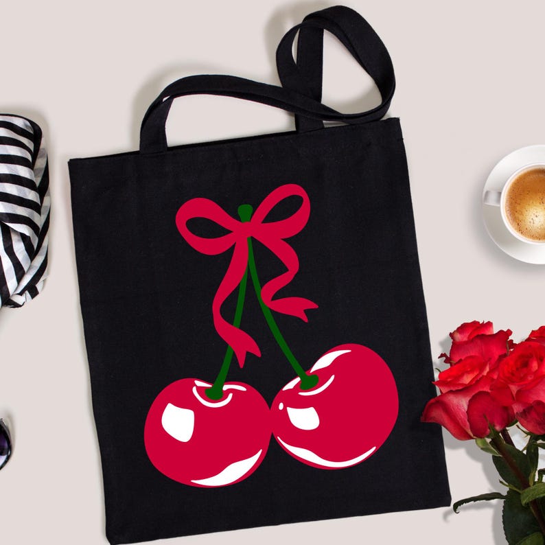 May include: Black tote bag featuring a red cherry design with a green stem and a red bow. The cherries have white highlights. The bag has two black handles.