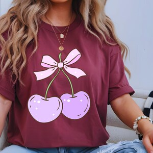 May include: Burgundy t-shirt featuring a cartoon design of two light purple cherries with green stems and a pink bow. The cherries have white highlights and a sparkly effect. The shirt has short sleeves.