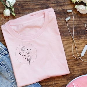 May include: A light pink t-shirt with a crew neck, featuring a black floral design inside a dotted circle. The design includes a flower, crescent moon, stars, and small dots. The shirt is laid on a wooden surface with jeans, flowers, and headphones.