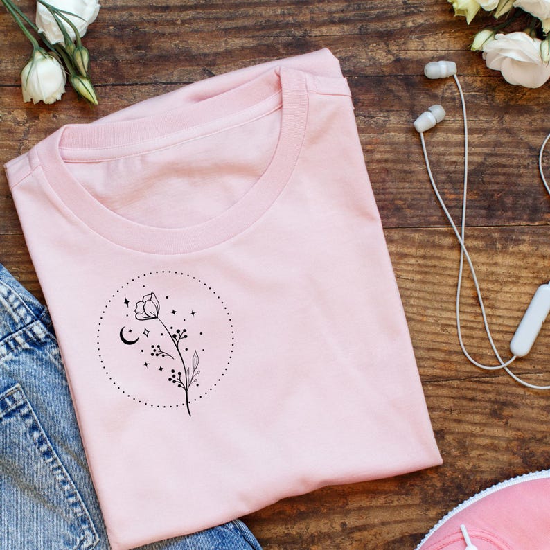 May include: A light pink t-shirt with a crew neck, featuring a black floral design inside a dotted circle. The design includes a flower, crescent moon, stars, and small dots. The shirt is laid on a wooden surface with jeans, flowers, and headphones.