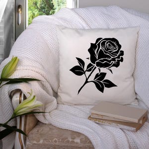 May include: A white square throw pillow featuring a large black rose design. The rose is detailed with petals and leaves. The pillow is placed on a white knitted blanket on a chair.