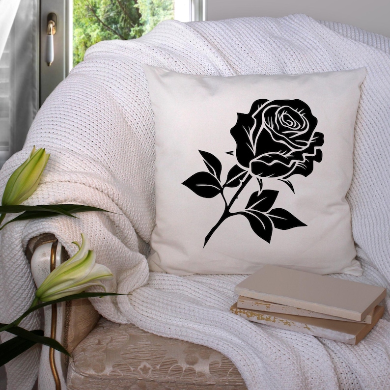 May include: A white square throw pillow featuring a large black rose design. The rose is detailed with petals and leaves. The pillow is placed on a white knitted blanket on a chair.