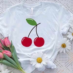 May include: White t-shirt featuring a graphic of two red cherries with a green leaf and black stems. The cherries have white highlights. The shirt is surrounded by pink tulips and white daisies.
