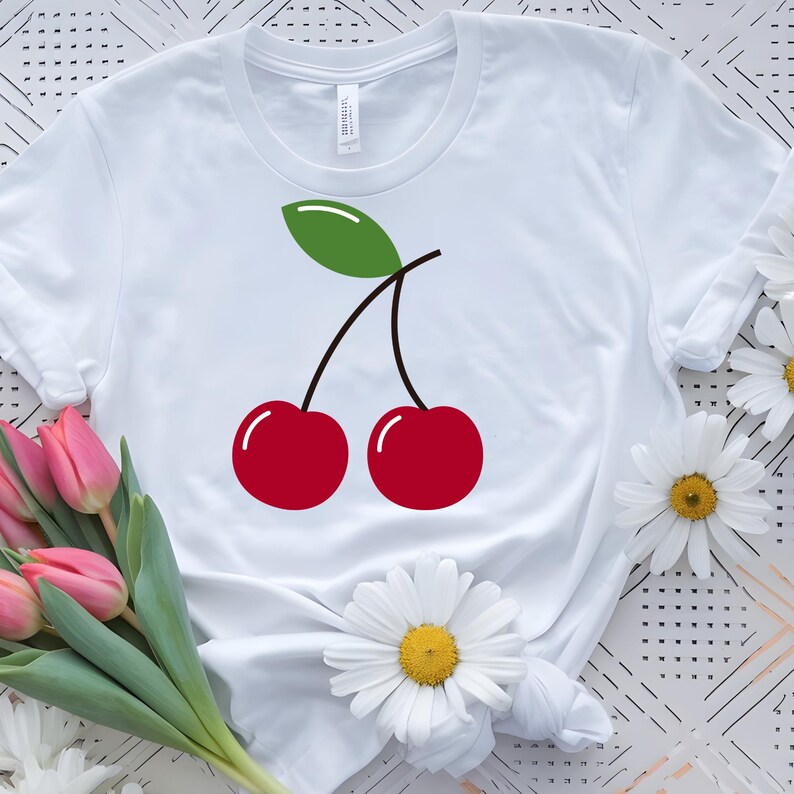 May include: White t-shirt featuring a graphic of two red cherries with a green leaf and black stems. The cherries have white highlights. The shirt is surrounded by pink tulips and white daisies.