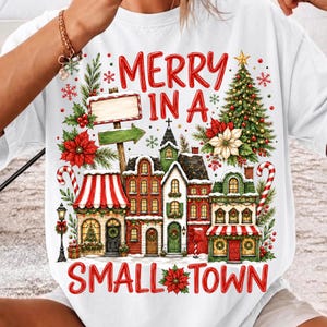 May include: A white t-shirt featuring a colorful graphic of a Christmas village with the text MERRY IN A SMALL TOWN. The design includes a decorated pine tree, red poinsettias, candy canes, a street lamp, and a wooden signpost on a snowy background.