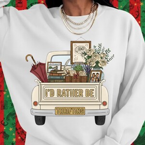 Puede incluir: Sudadera blanca con un diseño de caja de camión crema llena de libros, flores y cuadros enmarcados. El texto "I'D RATHER BE" está sobre una placa que dice "THRIFTING". Incluye un paraguas rojo.
