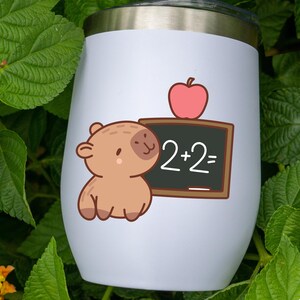 K&ouml;nnte beinhalten: Wei&szlig;er isolierter Becher mit einer Cartoon-Illustration eines braunen B&auml;ren neben einer Tafel mit der Gleichung "2+2=". Ein roter Apfel sitzt auf der Tafel. Der Becher hat einen silbernen Rand und Deckel.
