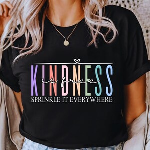 May include: Black t-shirt with the words "KINDNESS is free" in a colorful, rainbow-like font. The phrase "SPRINKLE IT EVERYWHERE" is below the main text. The shirt has short sleeves and a round neckline.