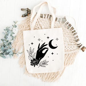 May include: Cream-colored tote bag with a black graphic of a hand holding a crescent moon, surrounded by stars and floral elements. The bag has cream-colored handles and is made of a sturdy fabric, perfect for carrying books or groceries.