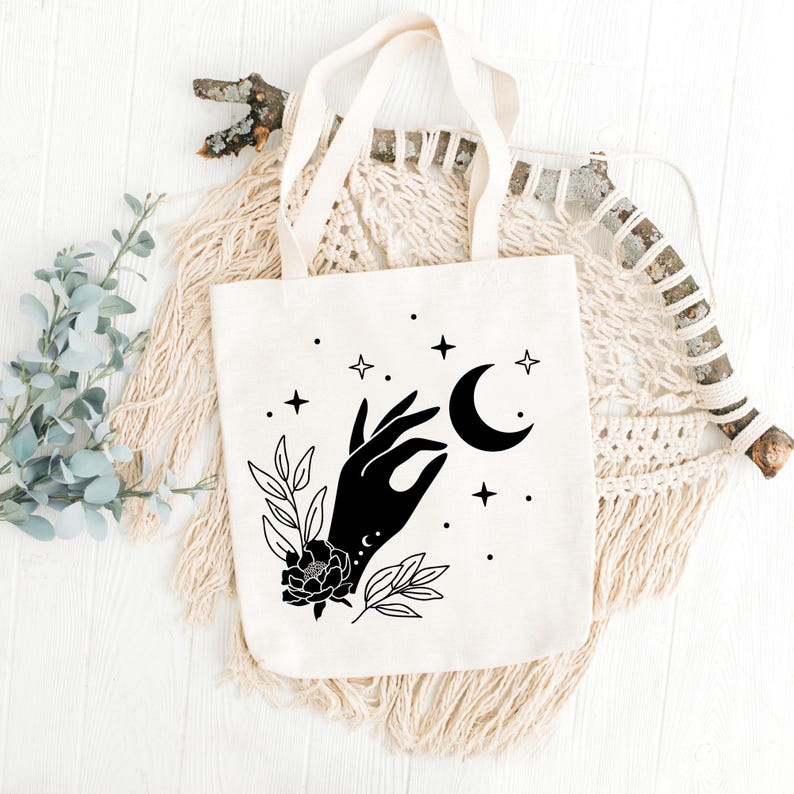 May include: Cream-colored tote bag with a black graphic of a hand holding a crescent moon, surrounded by stars and floral elements. The bag has cream-colored handles and is made of a sturdy fabric, perfect for carrying books or groceries.