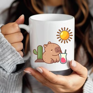 K&ouml;nnte beinhalten: Wei&szlig;e Keramik-Tasse mit einem Cartoon-Capybara, das aus einem Glas trinkt, einem gr&uuml;nen Kaktus und einer gelben Sonne. Die Tasse hat einen gro&szlig;en Henkel und wird von einer Person gehalten.