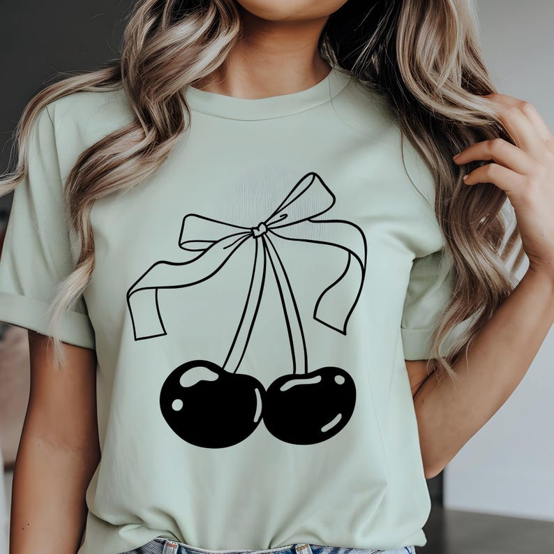 May include: A light green t-shirt featuring a black line drawing of two cherries tied together with a bow. The cherries have glossy highlights. The shirt has short sleeves and a crew neck.