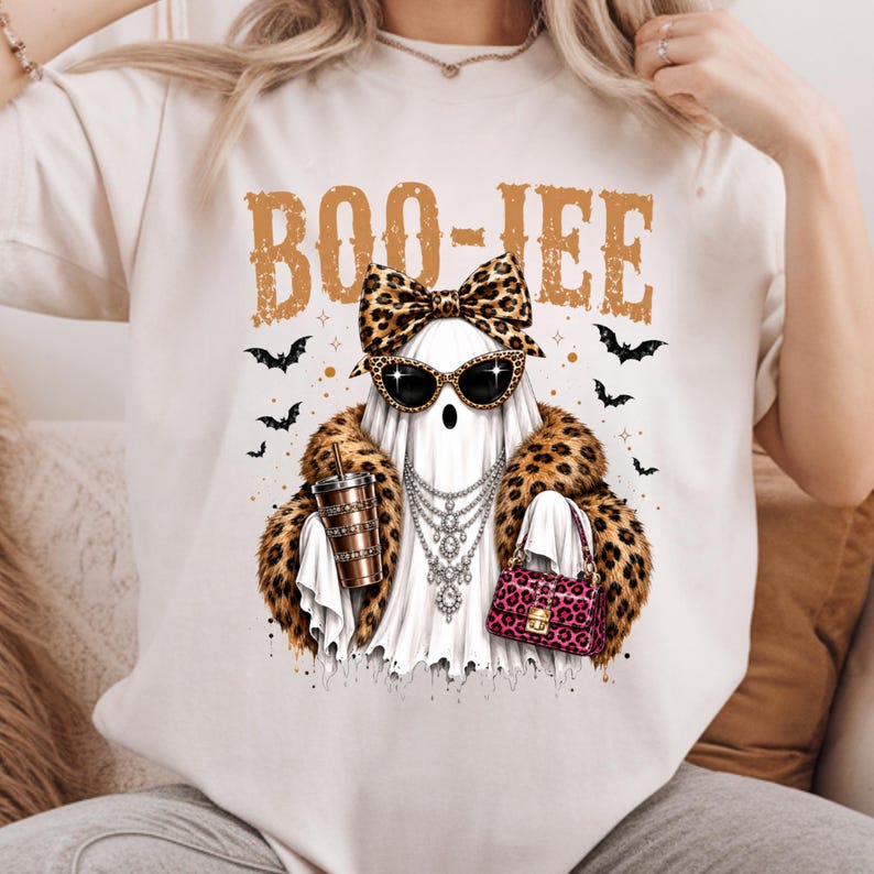 May include: A cream-colored t-shirt featuring a graphic of a ghost wearing a leopard print bow, cat-eye sunglasses, a layered silver necklace, and a leopard print fur coat. The ghost holds a tumbler and a pink handbag, with the text BOO-JEE above.