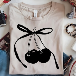 May include: Beige t-shirt featuring a black graphic of two cherries tied with a bow. The cherries are stylized with curved lines, and the bow adds a decorative element to the design. The shirt is made of a soft material.