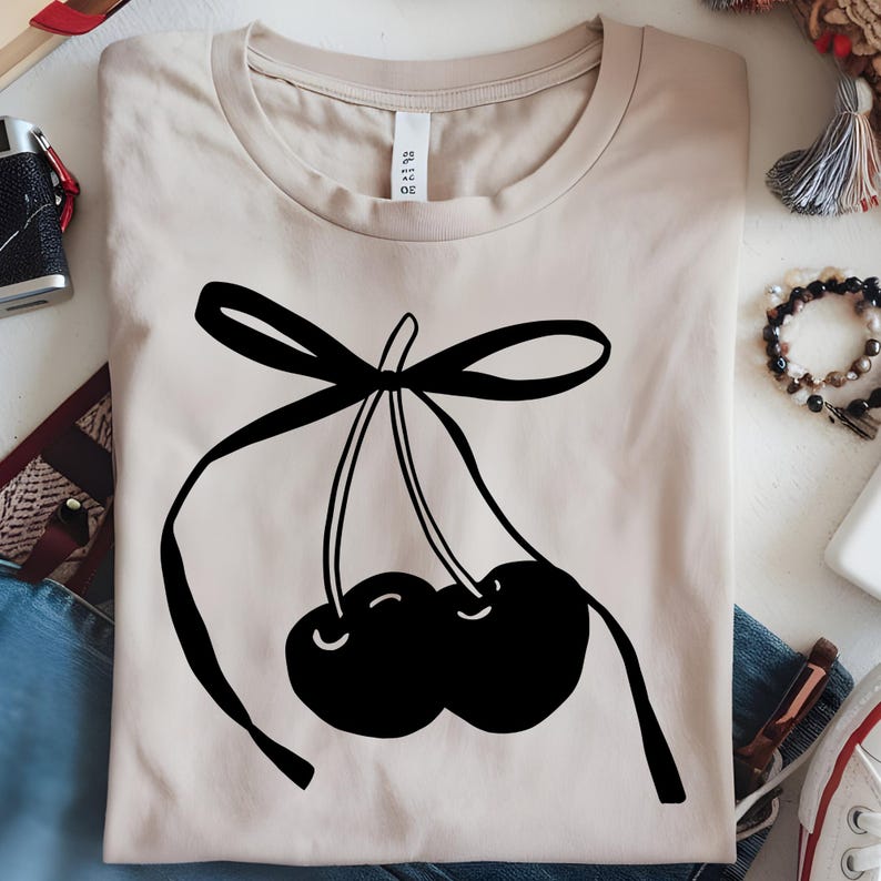 May include: Beige t-shirt featuring a black graphic of two cherries tied with a bow. The cherries are stylized with curved lines, and the bow adds a decorative element to the design. The shirt is made of a soft material.