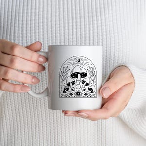 May include: White ceramic mug featuring a black and white illustration of mushrooms, a sun, and an eye. The design is framed by an arch with leaves. The mug has a curved handle.