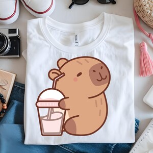 K&ouml;nnte beinhalten: Wei&szlig;es T-Shirt mit einem Cartoon-Capybara, das ein rosa Getr&auml;nk mit einem Strohhalm h&auml;lt. Das Capybara ist hellbraun mit einem l&auml;chelnden Gesicht. Das Getr&auml;nk befindet sich in einem transparenten Becher mit einem rosa Deckel. Das T-Shirt liegt auf einer wei&szlig;en Oberfl&auml;che.