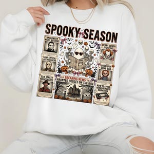 May include: A white long-sleeve sweatshirt featuring a graphic print with the text "SPOOKY SEASON" and "BREAKING NEWS: HAUNTED HOUSES ON THE RISE". The design includes illustrations of horror movie characters, skeletons, pumpkins, and a haunted house.