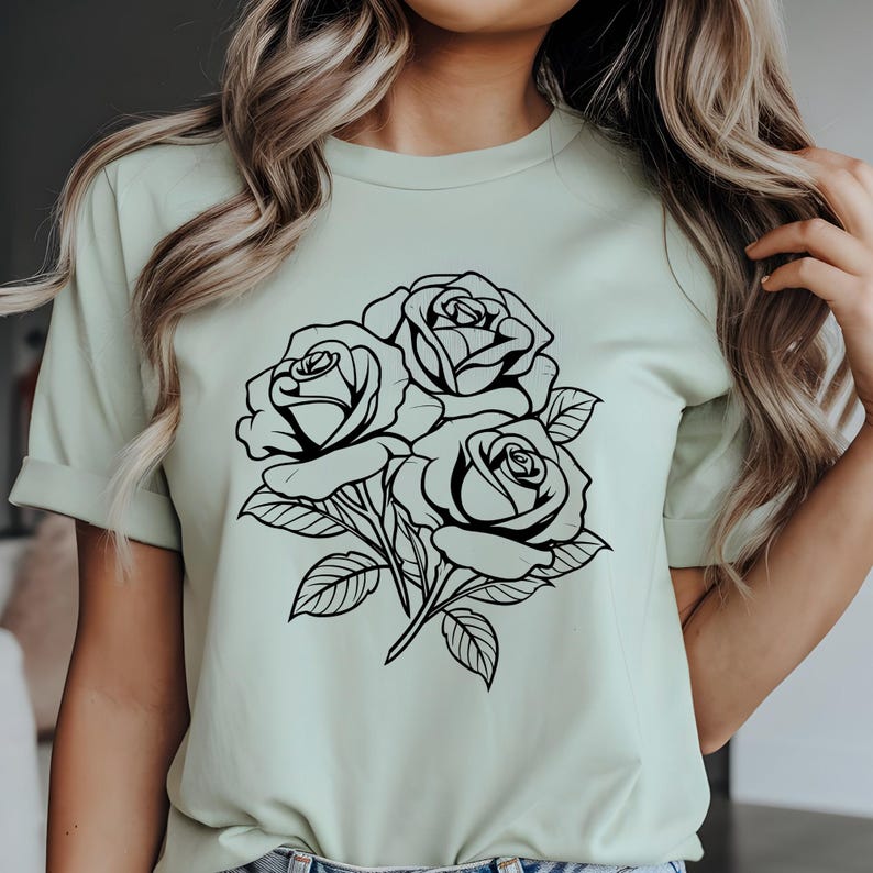 May include: A light green t-shirt featuring a black and white floral design of three roses with leaves. The roses are the focal point, with detailed outlines. The shirt has short sleeves and a crew neck.